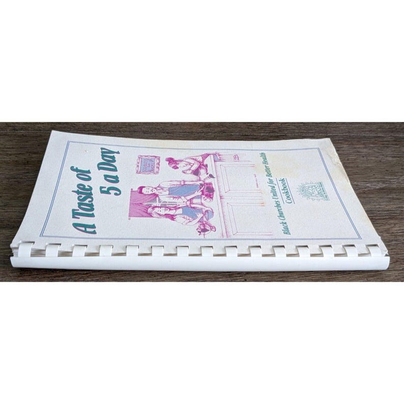 May include: A spiral-bound cookbook titled "A Taste of 5 a Day" with a vintage illustration on the cover. The cover features text in blue and a black and white illustration of people. The cookbook is open on a wooden surface.