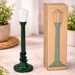May include: A miniature green street lamp with a white top, next to a cardboard box with a matching lamp illustration. The lamp has a black coiled detail near the base. The box has a vintage aesthetic.
