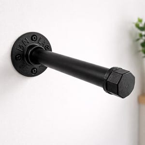 May include: Black industrial pipe towel rack. The rack features a round mounting plate with text and a straight pipe extending outward. The end of the pipe has a hexagonal cap. The rack is mounted on a white wall.