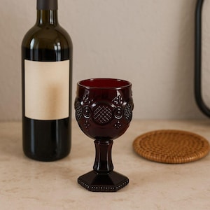 Könnte beinhalten: Ein weinrotes Weinglas mit Verzierungen, neben einer Weinflasche und einem gewebten Untersetzer. Das Glas hat einen sechseckigen Fuß und ein dekoratives Muster. Die Flasche hat ein leeres Etikett.
