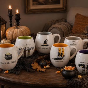 May include: A set of five white ceramic mugs with rounded bodies, each featuring unique Halloween-themed illustrations and text including Trick or Treat, Wicked Brew, Feeling Wicked, Have a Bewitching, and Smell My Feet, with colorful interior glazes.