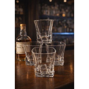 May include: Four clear, square-shaped whiskey glasses with a textured design, one stacked on top of the others. A bottle of whiskey with a cork stopper is in the background, suggesting a bar setting. The glasses are suitable for spirits.