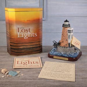 May include: A Lefton Lost Lights lighthouse figurine on a wooden base, accompanied by a sunset-themed metal tin, a certificate of authenticity, and a descriptive card. The lighthouse features a brick-patterned tower and a rocky base.