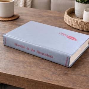 Op de afbeelding: Een hardcover boek getiteld "Sheridan in the Shenandoah" met een lichtblauwe kaft. Het boek heeft een rode illustratie en tekst. Het boek ligt op een houten oppervlak.