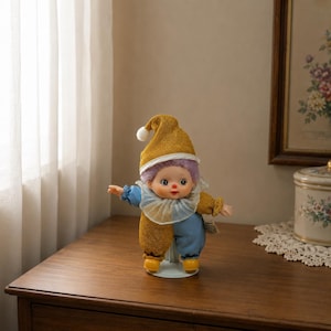 May include: A small doll dressed as a jester, featuring a gold and blue fabric outfit with a matching gold pointed hat topped with a white pom-pom. The doll has light purple hair, a ruffled collar, and yellow shoes, standing on a circular base.