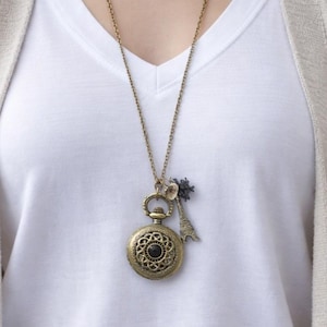 Peut inclure: Un long collier avec un pendentif montre de poche rond en laiton antique, orné d'un motif filigrane complexe et d'une pierre noire centrale. Deux petites breloques sont attachées au pendentif : une tour Eiffel et une fleur foncée.