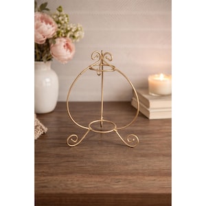 May include: A gold-colored metal display stand with a heart-shaped design. The stand has a circular base and three decorative feet. A small candle and a vase of flowers are in the background.