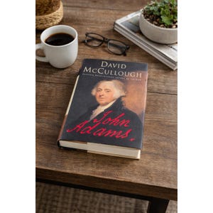May include: A hardback book titled John Adams by David McCullough featuring a portrait of John Adams on the cover. The book is positioned near a white ceramic mug containing black coffee, a pair of black-rimmed spectacles, and a small potted plant.