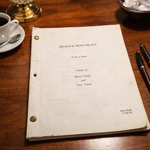 May include: A script for "3rd Rock From The Sun" titled "Truth or Dick" by Bonnie and Terry Turner. The first draft is dated 7/28/95. The script is on a wooden table with a cup of coffee, a spoon, and two pens.