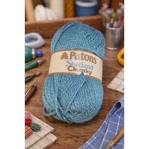 May include: A ball of teal-colored Patons Shetland Chunky yarn with a brown paper label. The label features the brand name and yarn type. Knitting needles and other craft supplies are in the background.