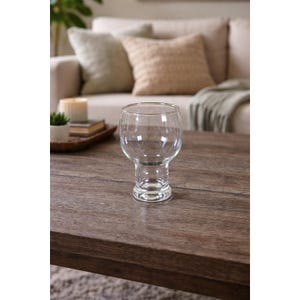 May include: A clear, stemmed glass with a rounded bowl and a short, sturdy base. The glass sits on a dark brown wooden table, with a blurred background of a beige sofa and decorative pillows. The glass is empty.
