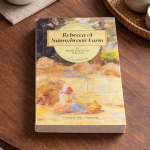 Puede incluir: Un libro de bolsillo titulado Rebecca of Sunnybrook Farm de Kate Douglas Wiggin, parte de la serie Wordsworth Classics. La portada presenta una ilustración de una persona con un gorro rosa sentada en un campo, con el texto Complete and Unabridged.