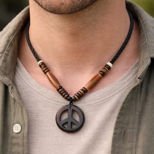 May include: A wooden peace sign pendant necklace. The necklace features a dark brown peace symbol pendant, wooden beads, and a black cord. The necklace is worn against a neutral background.