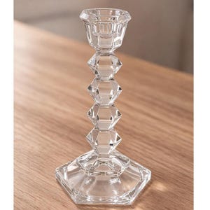 May include: A clear glass candlestick holder with a faceted, geometric design. The base is a hexagonal shape, and the stem features stacked diamond shapes. The top has a small, round cup to hold a candle. The candlestick is made of clear glass.