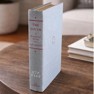 May include: A light blue book titled "THE SOUTH" with red text and star accents. The book is a documentary history by Van Noppen, published by Van Nostrand. The book is standing upright on a wooden surface.