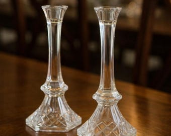 May include: Two clear glass candlesticks. Each candlestick has a tall, slender stem and a hexagonal base with a diamond pattern. The top of each candlestick is designed to hold a candle. The candlesticks are displayed on a wooden surface.