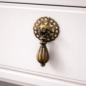 以下が含まれることがあります： 真鍮製の装飾的な引き出しの取っ手のクローズアップ。取っ手は、サンバーストのデザインの装飾的な円形のバックプレートと、涙の形をしたハンドルを備えています。ハードウェアは白い表面に取り付けられています。