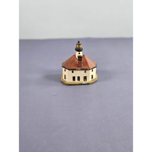 Può includere: Modello di faro in miniatura con base bianca, tetto marrone e torre nera e oro. Il faro ha piccole finestre rettangolari e poggia su una base beige. Un oggetto decorativo.
