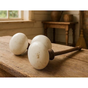 May include: Three off-white ceramic cabinet knobs with dark brown wooden stems rest on a wooden surface. The knobs have a speckled appearance. The background includes a window, a small table, and a broom, suggesting a rustic or farmhouse style.