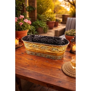 以下が含まれることがあります： 金色の仕上げと複雑な渦巻き模様が施された、装飾的な長方形のプランター。 プランターは黒い内側で、木製のテーブルの上に置かれています。 デザインには緑色のアクセントが含まれています。 背景には鉢植えの植物と屋外の座席があります。