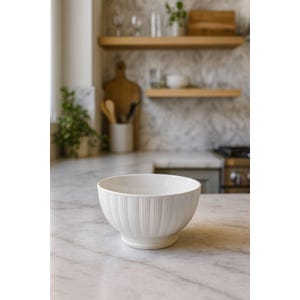 May include: A white ceramic bowl with vertical ribbed detailing. The bowl has a wide opening and a rounded base. It is sitting on a marble countertop in a kitchen setting. The background includes wooden shelves and a stove.