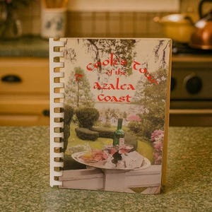 May include: A spiral-bound cookbook titled "Cooks of the Azalea Coast" with a vintage cover featuring a scenic outdoor image. The cover art includes a bottle of wine, glasses, and a tray of food. The book is on a green countertop.