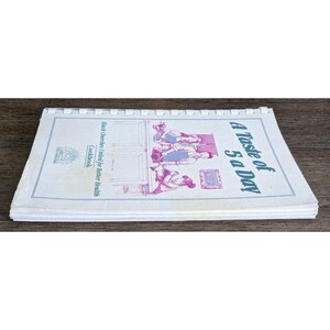 May include: Open cookbook titled "A Taste of 5 a Day" with a spiral binding. The cover features illustrations in pink and blue, with text that reads "Black Churches United for Better Health Cookbook."