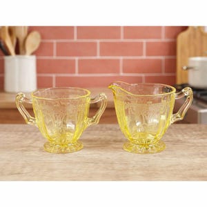 May include: A pair of yellow pressed glass vessels consisting of a two-handled sugar bowl and a creamer pitcher. Both pieces feature a detailed embossed pattern of swags and garlands, a scalloped rim, and a circular pedestal base.