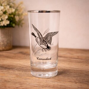 May include: A clear glass tumbler featuring a black ink illustration of a flying duck with the text Canvasback printed below the bird. The glass has a silver-colored rim and a thick, weighted base, providing a sturdy structure for this drinkware.