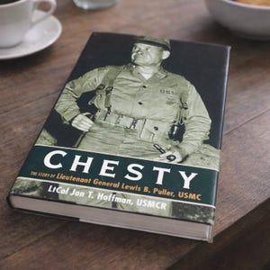 May include: A hardcover book titled "CHESTY" on a wooden surface. The book cover features a black and white image of a military officer in uniform. The title is in large white letters with a green banner.
