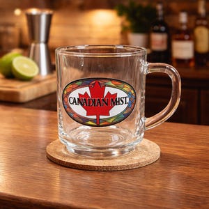 May include: A clear glass mug with a handle, featuring an oval sticker with a red maple leaf and the words "CANADIAN MIST." The mug sits on a round cork coaster. The background includes bar tools and bottles.
