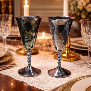 May include: Two silver goblets with twisted stems sit on a table set for a formal dinner. The goblets have a flared bowl and a detailed, textured stem. Lit candles and other glassware are in the background, creating a warm, inviting atmosphere.