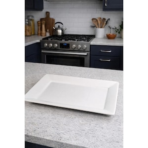 May include: A rectangular, white serving platter with slightly raised edges. The platter is made of a glossy material and sits on a speckled countertop in a kitchen setting. The kitchen features dark blue cabinets and a stainless steel stove.