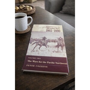 Op de afbeelding: Een hardcover boek met de titel "Eyewitnesses to the Indian Wars 1865-1890" ligt open op een donkere houten tafel. De kaft toont een sepia-kleurige illustratie van soldaten te paard. Het boek is Volume Two, "The Wars for the Pacific Northwest" door Peter Cozzens.