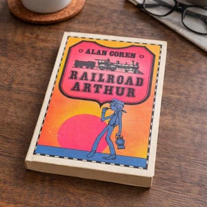 Puede incluir: Un libro vintage titulado "Railroad Arthur" de Alan Coren. La portada presenta una colorida ilustración de un tren, una figura con una linterna y una puesta de sol rosa y naranja. El libro está sobre una superficie de madera.