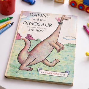 Puede incluir: Un libro infantil vintage titulado "Danny and the Dinosaur" de Syd Hoff. La portada presenta una ilustración de dibujos animados de un dinosaurio con un niño en su cuello. El libro es un "I CAN READ Book". Crayones y juguetes rodean el libro.