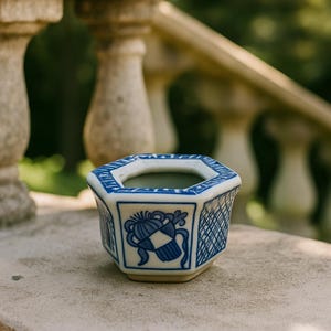 May include: A small, octagonal ceramic planter with a white base and blue decorative patterns. The planter features a blue and white design on each side, with a detailed floral motif and a lattice pattern. The interior is a light green colour.