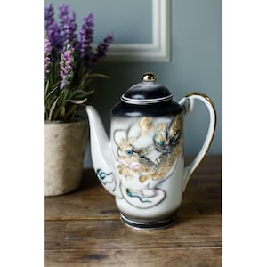 Op de afbeelding: Een wit en zwarte keramische theepot met een gouden drakenontwerp. De theepot heeft een gouden handvat en een gouden knop op het deksel. De draak is gedetailleerd met gouden, blauwe en turquoise accenten. Een decoratief stuk voor theeliefhebbers.