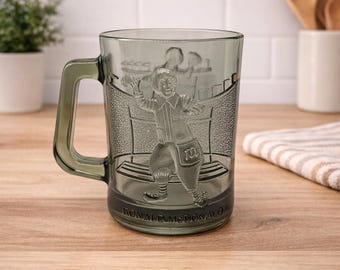 May include: A grey glass mug with a handle, featuring an embossed image of Ronald McDonald. The mug has the words "Ronald McDonald" at the base. The mug is on a wooden surface.