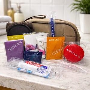Könnte beinhalten: Ein Reise-Set mit verschiedenen Toilettenartikeln, darunter eine braune Kulturtasche, ein schwarzes Gesichtspad und Päckchen mit der Aufschrift "FRESHEN" und "CLEAN". Weitere Artikel sind eine Zahnbürste, Zahnpasta und ein roter runder Artikel mit "DELTA".