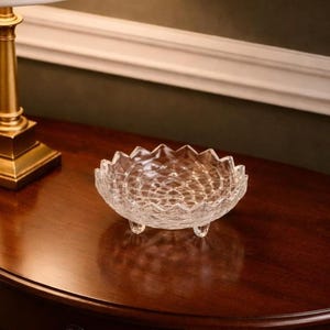 Puede incluir: Un cuenco de cristal transparente, pequeño y decorativo, con borde festoneado y tres patas pequeñas. El cuenco tiene un patrón texturizado en forma de diamante. El cuenco está sobre una superficie de madera marrón oscuro.