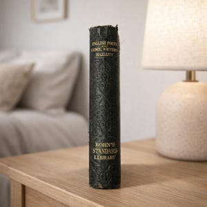 May include: A vintage book with a dark green cover and gold lettering. The book's title is "English Poets and Comic Writers" and "Bohn's Standard Library". The book is standing upright on a wooden surface.