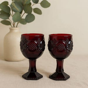 Puede incluir: Dos copas de vino tinto rubí con detalles en relieve ornamentados. Las copas tienen un tallo corto y una base cuadrada. El fondo incluye un jarrón color crema con hojas verdes.