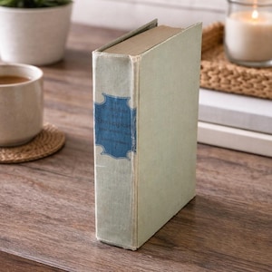 May include: A vintage book with a light green cover and a blue label that reads "Complete Shakespeare". The book is standing upright on a wooden surface, with a cup of coffee and a small plant in the background.