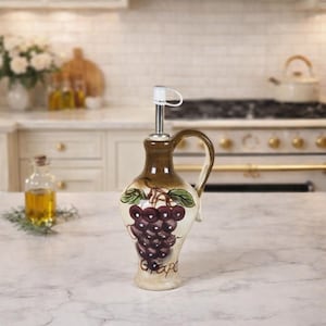 May include: A ceramic olive oil dispenser with a brown handle and spout. The dispenser features a painted design of purple grapes and green leaves. The word "Grape" is written at the bottom of the dispenser.