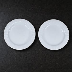 Puede incluir: Dos platos de cerámica blanca con un diseño de borde acanalado sobre una superficie oscura. Los platos son redondos y tienen una estética simple y elegante, adecuada para diversos entornos de comedor.