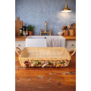 May include: Rectangular woven basket with rope handles, decorated with ceramic vegetables. The basket is made of light brown woven material and sits on a wooden surface. The background features a kitchen sink and faucet.