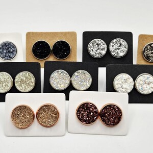 May include: A collection of stud earrings in various colors and textures. The earrings are round and feature a sparkly, druzy-like appearance. They are displayed on white, black, and brown earring cards. Colors include silver, black, gold, and shades of pink.