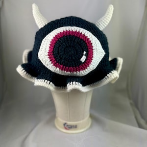 May include: A handmade crochet hat in navy blue, featuring a large white and magenta eye design. The hat has two white horns and a ruffled white brim. The hat is displayed on a white mannequin head.