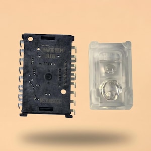 May include: A rectangular black electronic component with multiple metal pins along its sides and printed text reading BMW36 100M and NS398932C, positioned next to a clear, rectangular plastic housing containing a circular lens element.